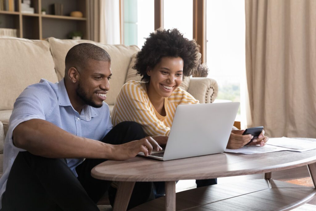 A couple sits on the floor to pay bills on their laptop and are smiling.